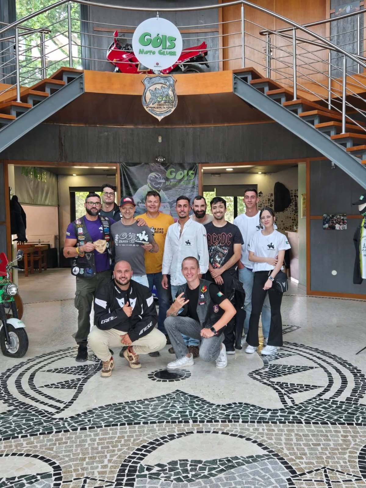 Fotografia de grupo na sede de um motoclube, com emblema e uma mota vermelha ao fundo