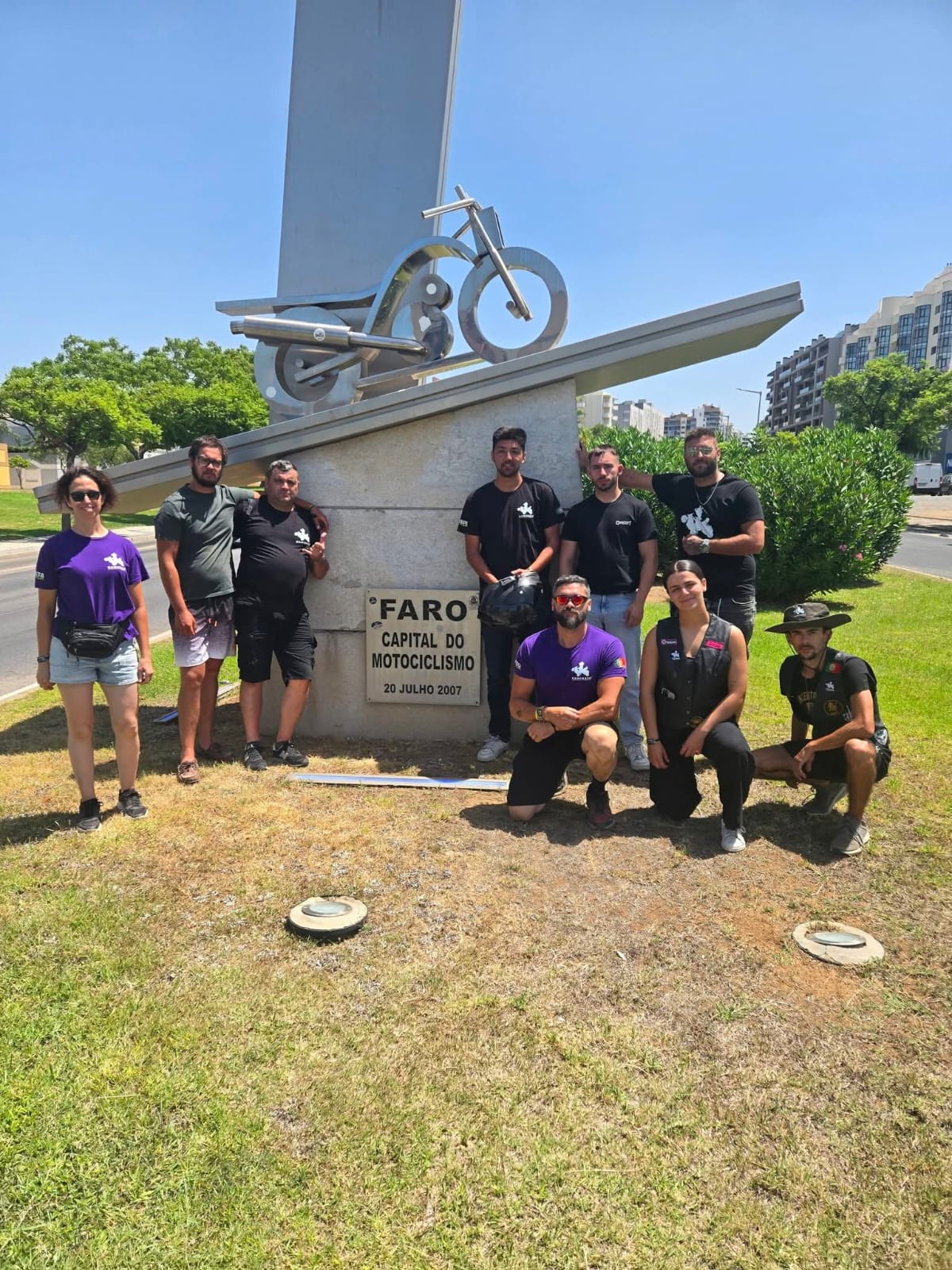 Grupo de membros do clube junto a um monumento de mota em aço, com a placa Faro Capital do Motociclismo