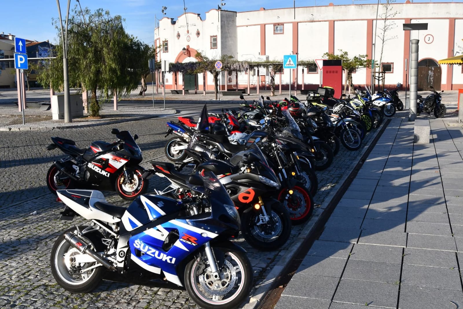 Várias motas estacionadas num largo empedrado em frente a uma praça de touros