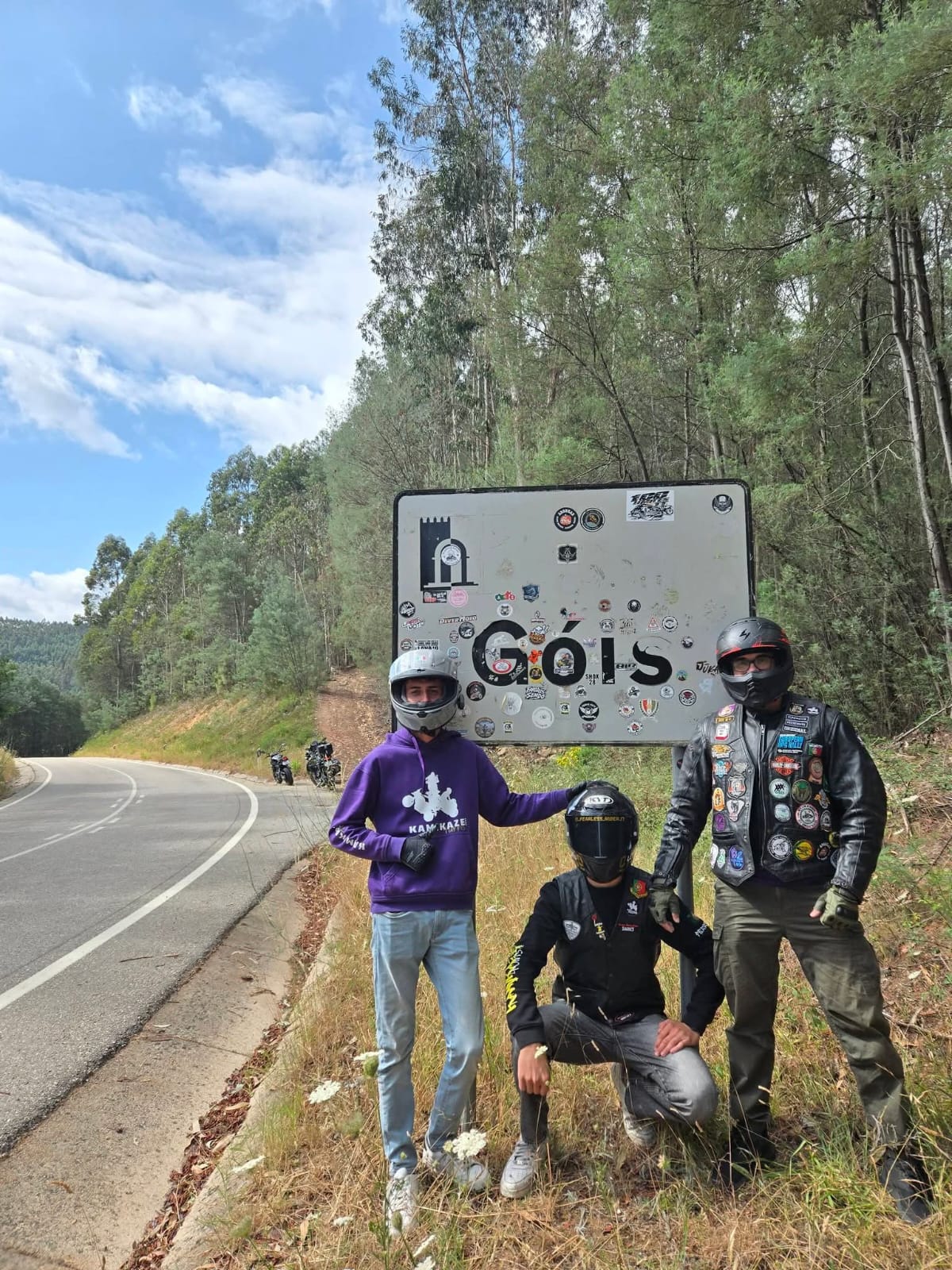 Três membros do clube junto ao painel de entrada de Góis, com motas ao fundo na estrada