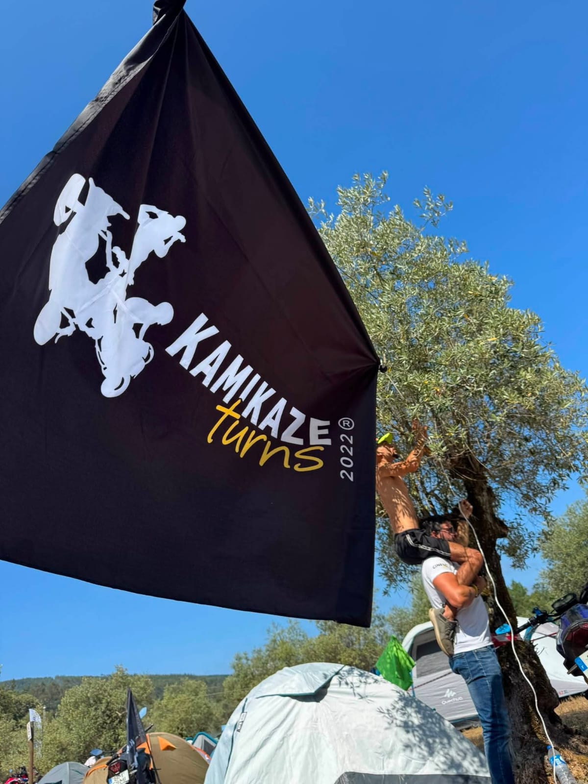 Bandeira preta do clube ao vento num campo de campismo, com dois membros ao fundo
