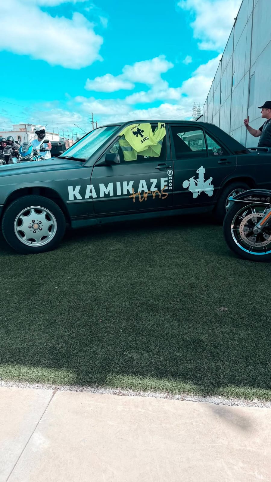 Roda dianteira de uma mota preta junto a um automóvel decorado com a marca Kamikaze Turns