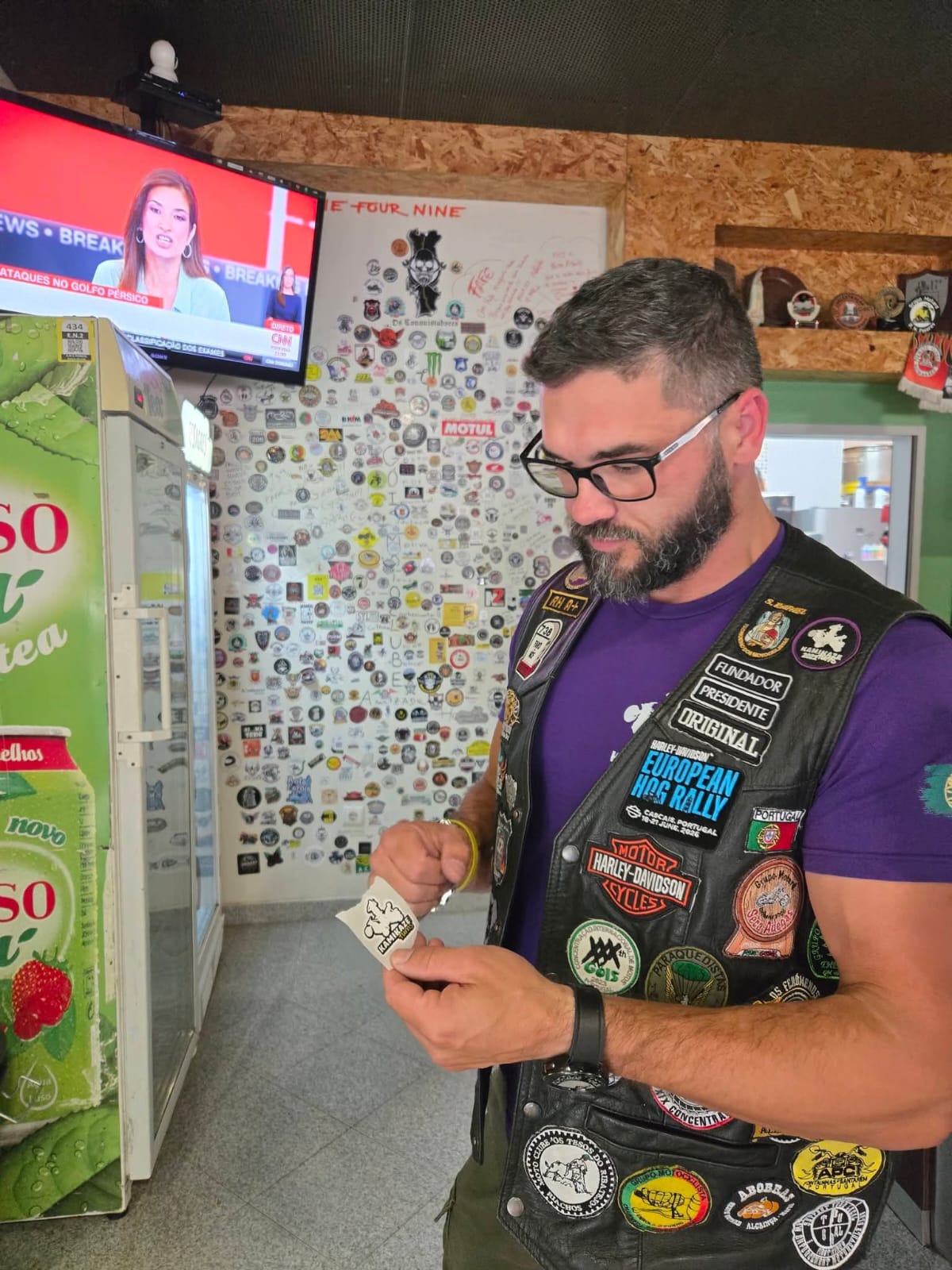Membro do clube, de colete bordado, a segurar um patch Kamikaze numa sala coberta de emblemas