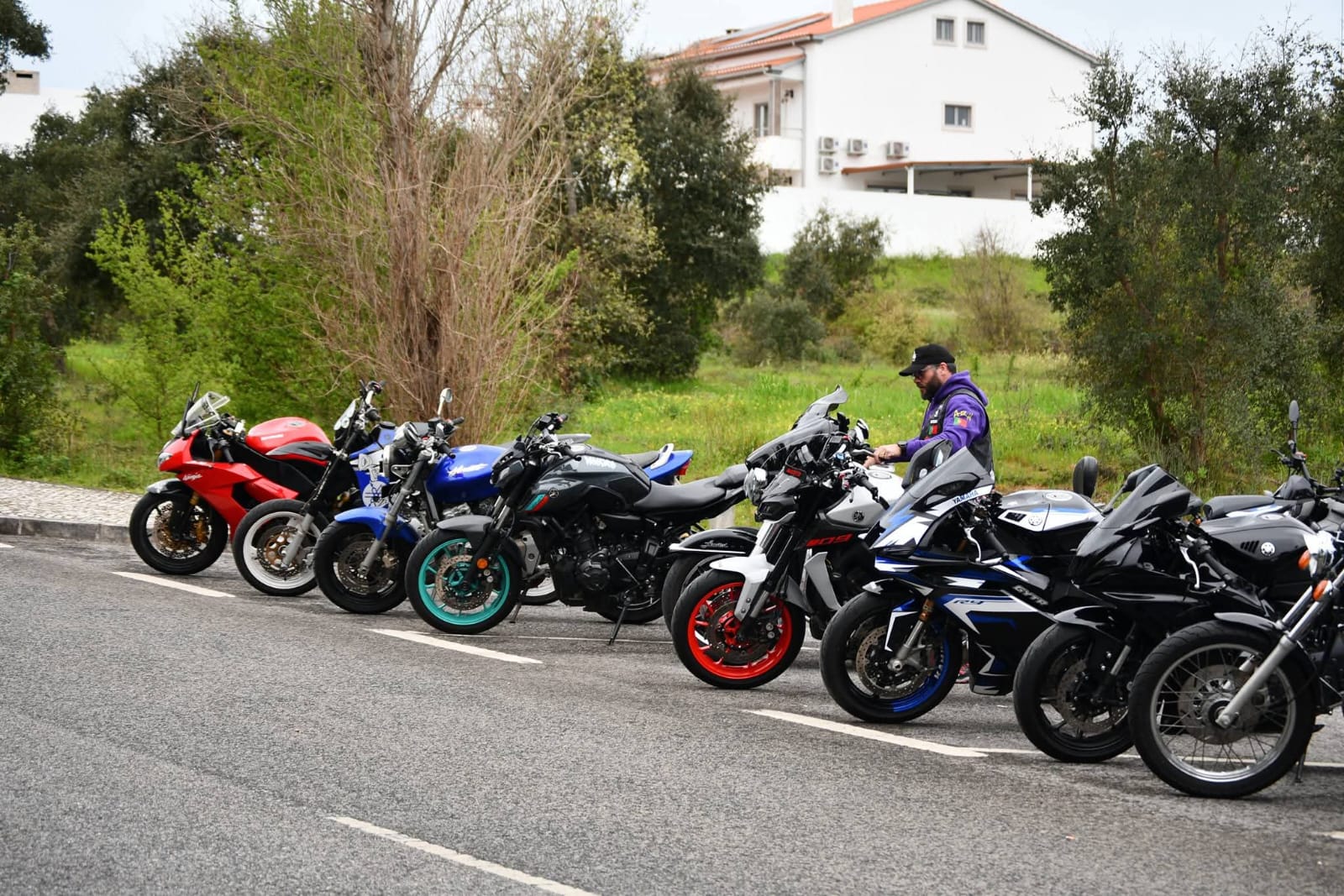 Motas alinhadas num parque de estacionamento, com um motociclista de casaco roxo do clube