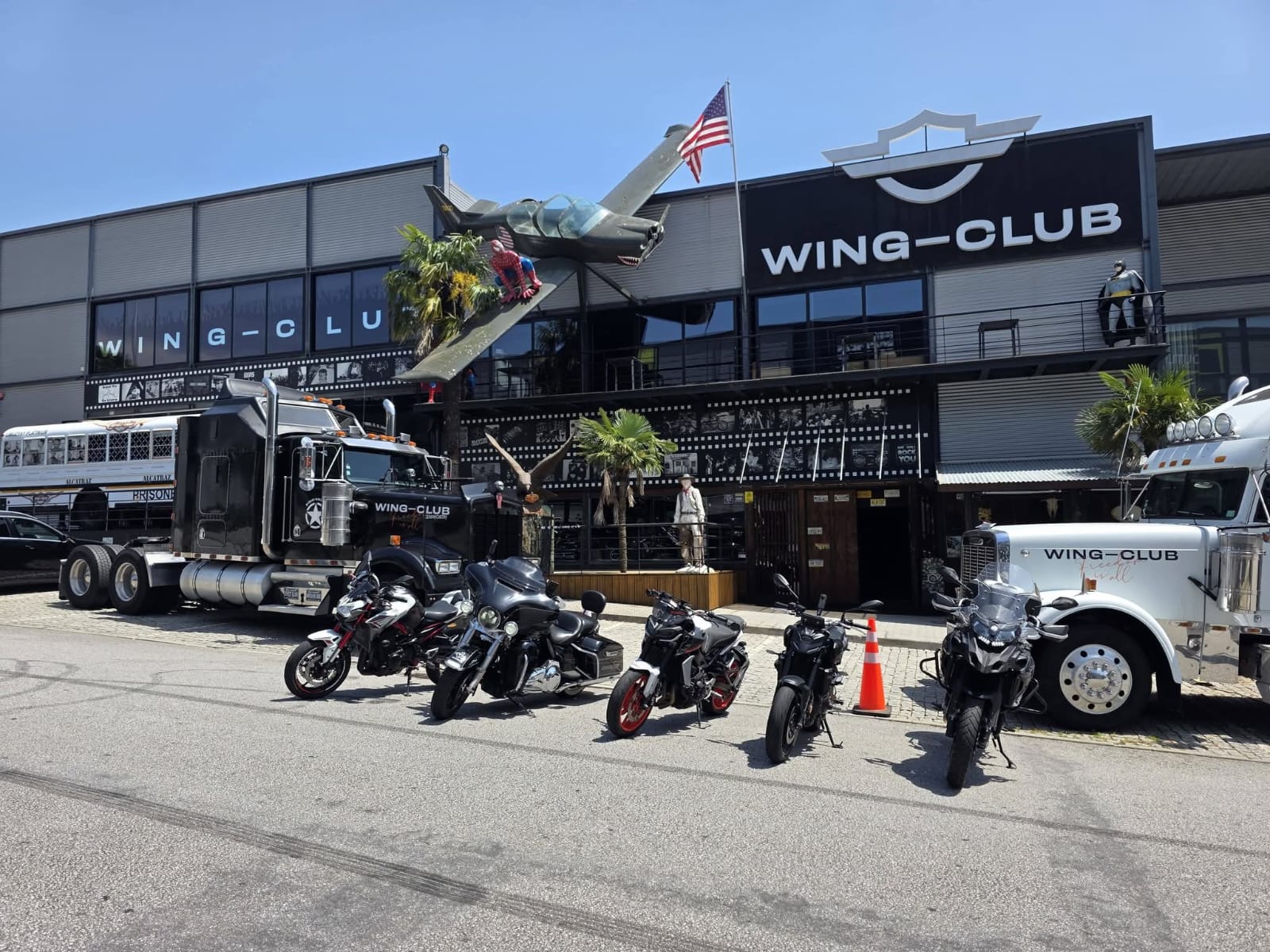 Motas estacionadas junto a um edifício com a inscrição Wing-Club e veículos americanos